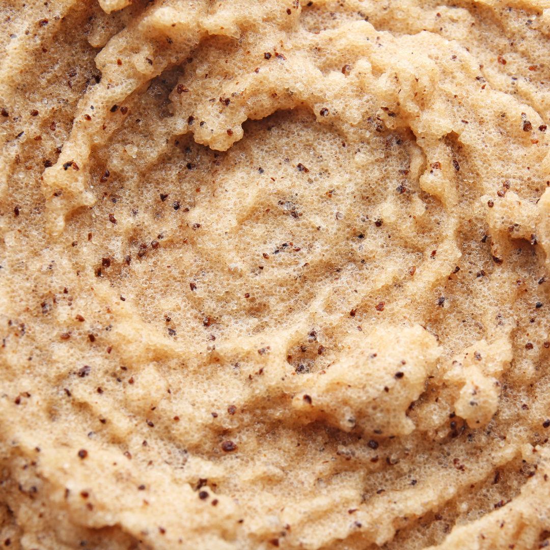 Close-up image of Bumbee Beauty Coffee Scrub. The texture is creamy and slightly coarse, with visible coffee grounds dispersed throughout the beige-colored scrub. The consistency looks rich and exfoliating, highlighting the natural ingredients used in the product.
