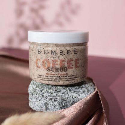 A jar of Bumbee Beauty Coffee Scrub placed on a rock with a backdrop of a soft pink colour. The jar has a white lid and a label that reads 'Refresh + Hydrate,' listing ingredients like coffee, coconut, shea butter, and vitamin E. The natural and floral setting emphasizes the product's connection to nature and beauty