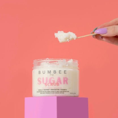 A hand scooping up a spoon full of sugar scrub from a Bumbee Beauty Sugar Scrub 8oz jar, revealing its beautiful and exfoliating texture. Bumbee Beauty Sugar scrub highlights ingredients such as coconut oil and Shea Butter promising gentle exfoliation and nourishment for the skin.